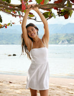 Woman in a white dress standing on a beach with trees and mountains in the background.