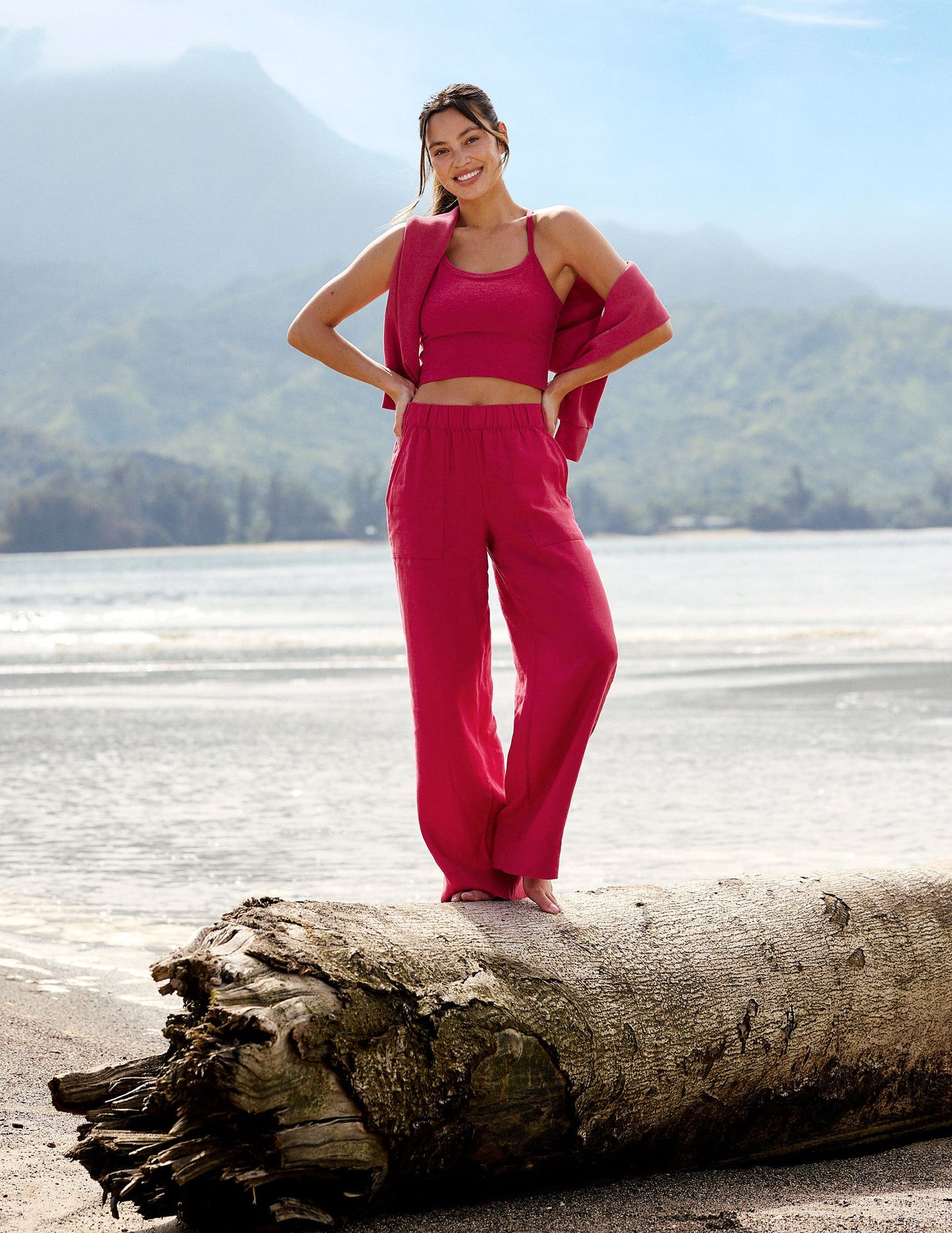 Woman wearing a red cropped tank and linen pants standing on a tree branch in front of the ocean.