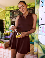 Woman in a brown sleeveless top and shorts holding a small papaya in a tropical-themed setting.