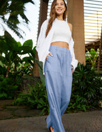Woman wearing a white top and blue pants standing outdoors with greenery in the background.
