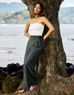 Woman in a white top and dark pants standing next to a tree by a body of water with mountains in the background.