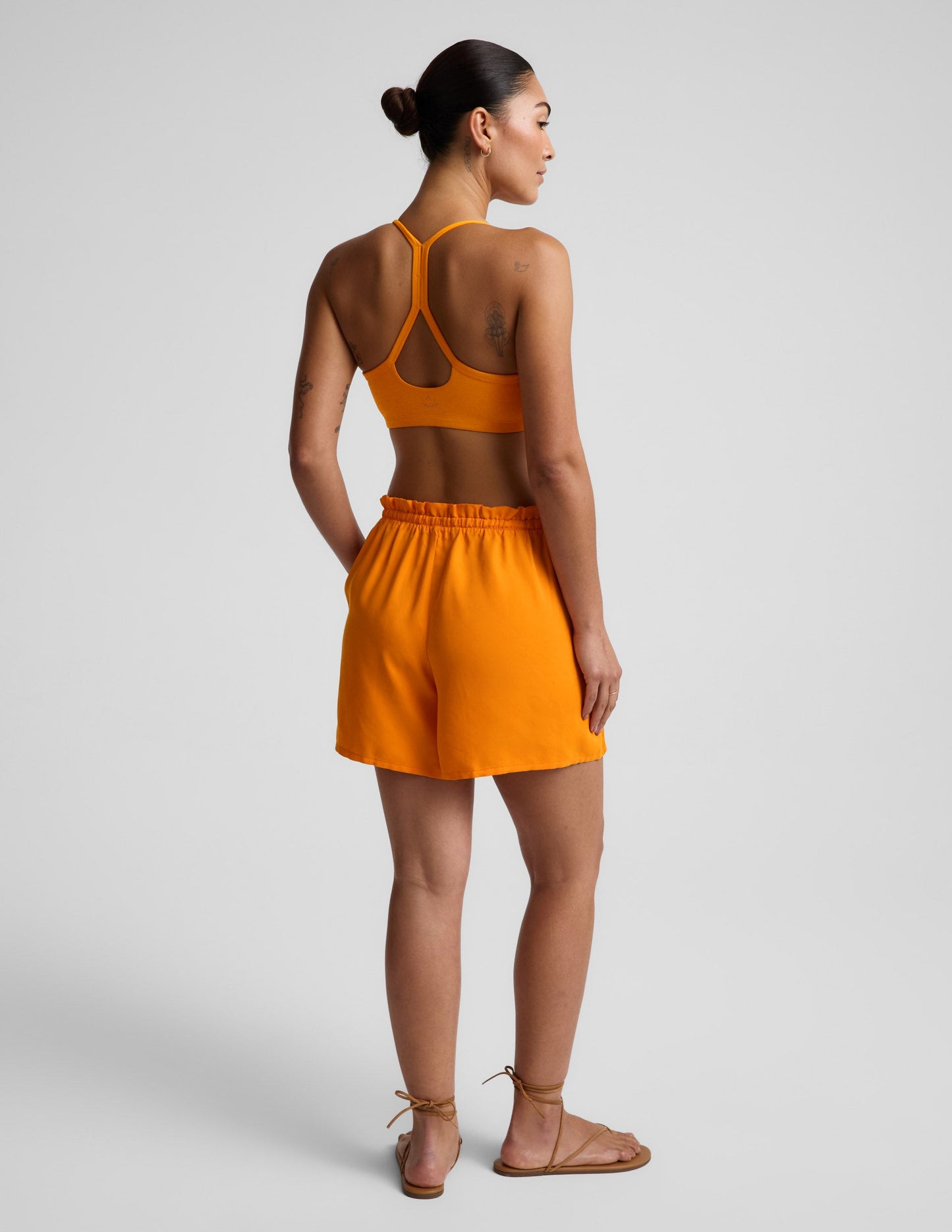 Woman wearing orange SunKissed Cupro Drawstring Short in a rear view, paired with a matching sports bra and sandals.