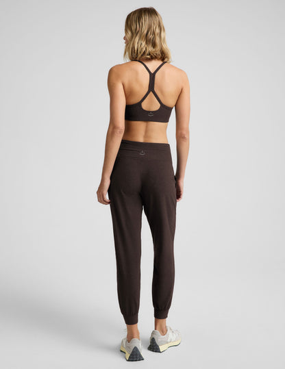 A woman viewed from the back wearing a dark brown sports bra and matching jogger-style leggings with white sneakers, photographed against a light gray studio background.