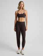 A woman modeling a matching dark brown activewear set consisting of a sports bra and jogger pants, paired with white sneakers, against a light gray studio background.