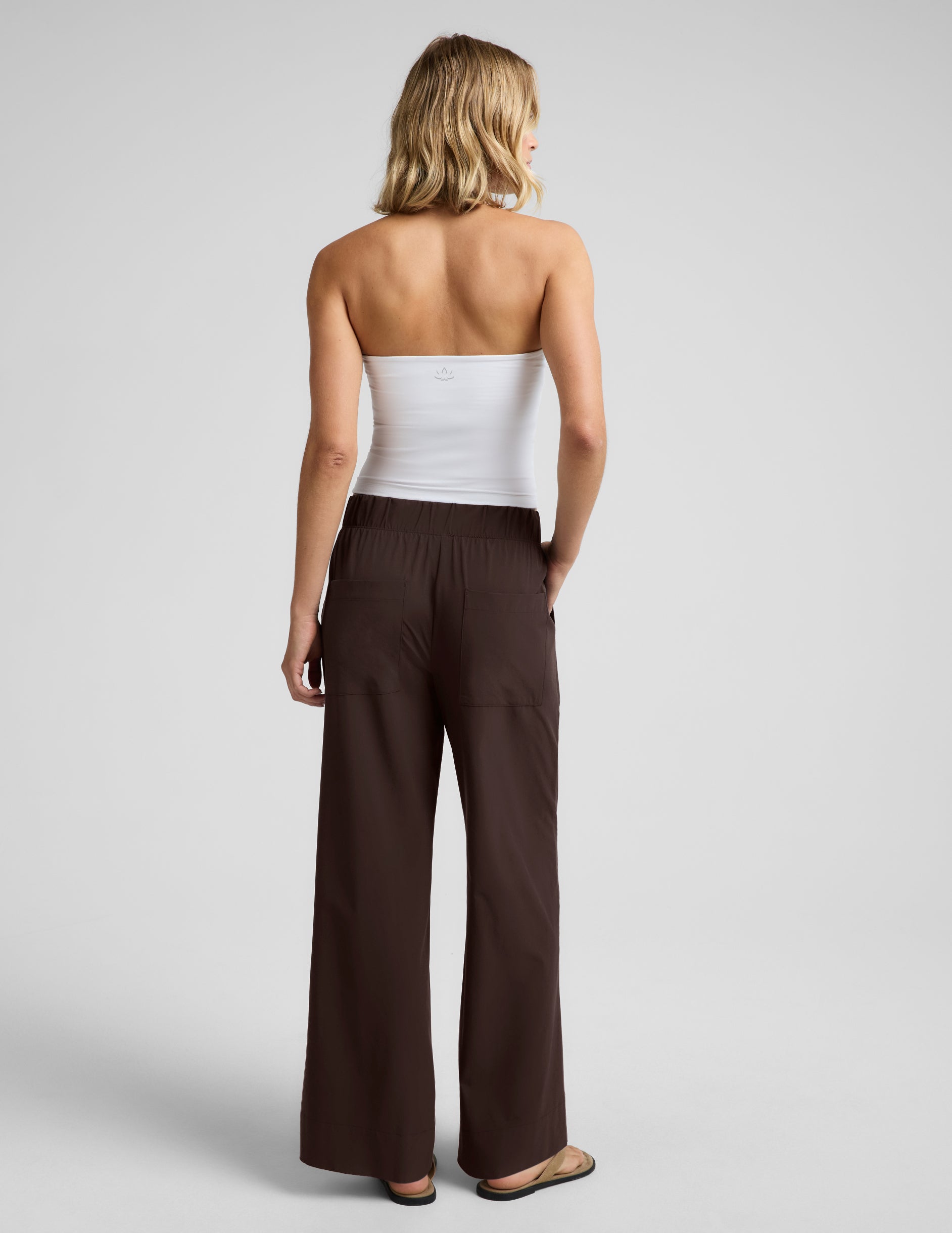 Woman wearing a white halter tank top and brown wide leg pants against a plain studio background.