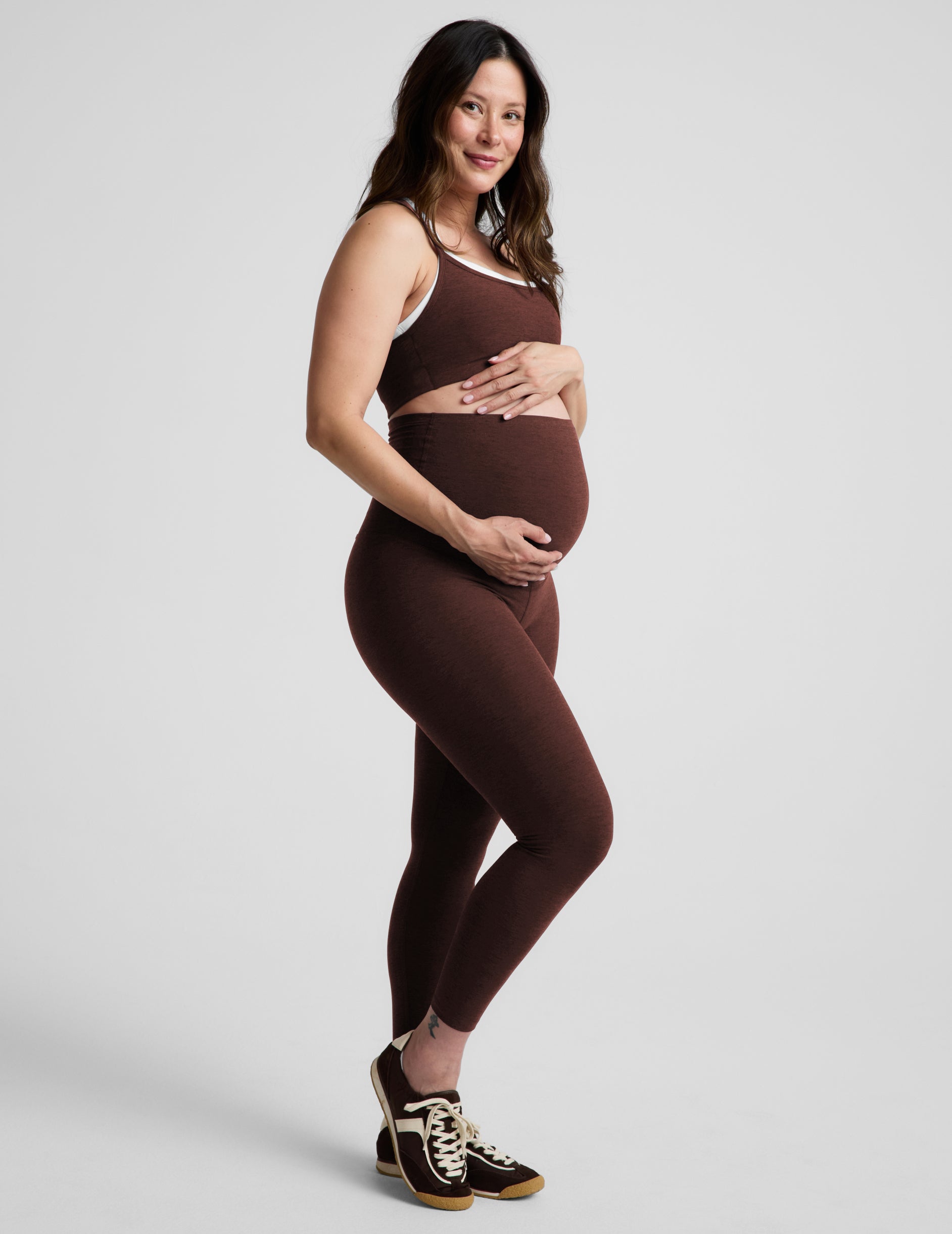 Pregnant woman wearing a brown sports bra and brown maternity leggings against a plain background.