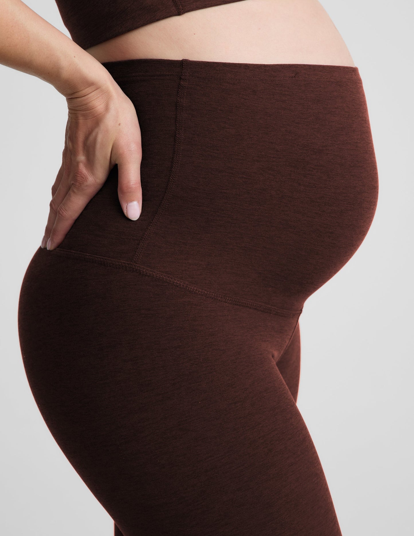 Pregnant woman wearing a brown sports bra and brown maternity leggings against a plain background.