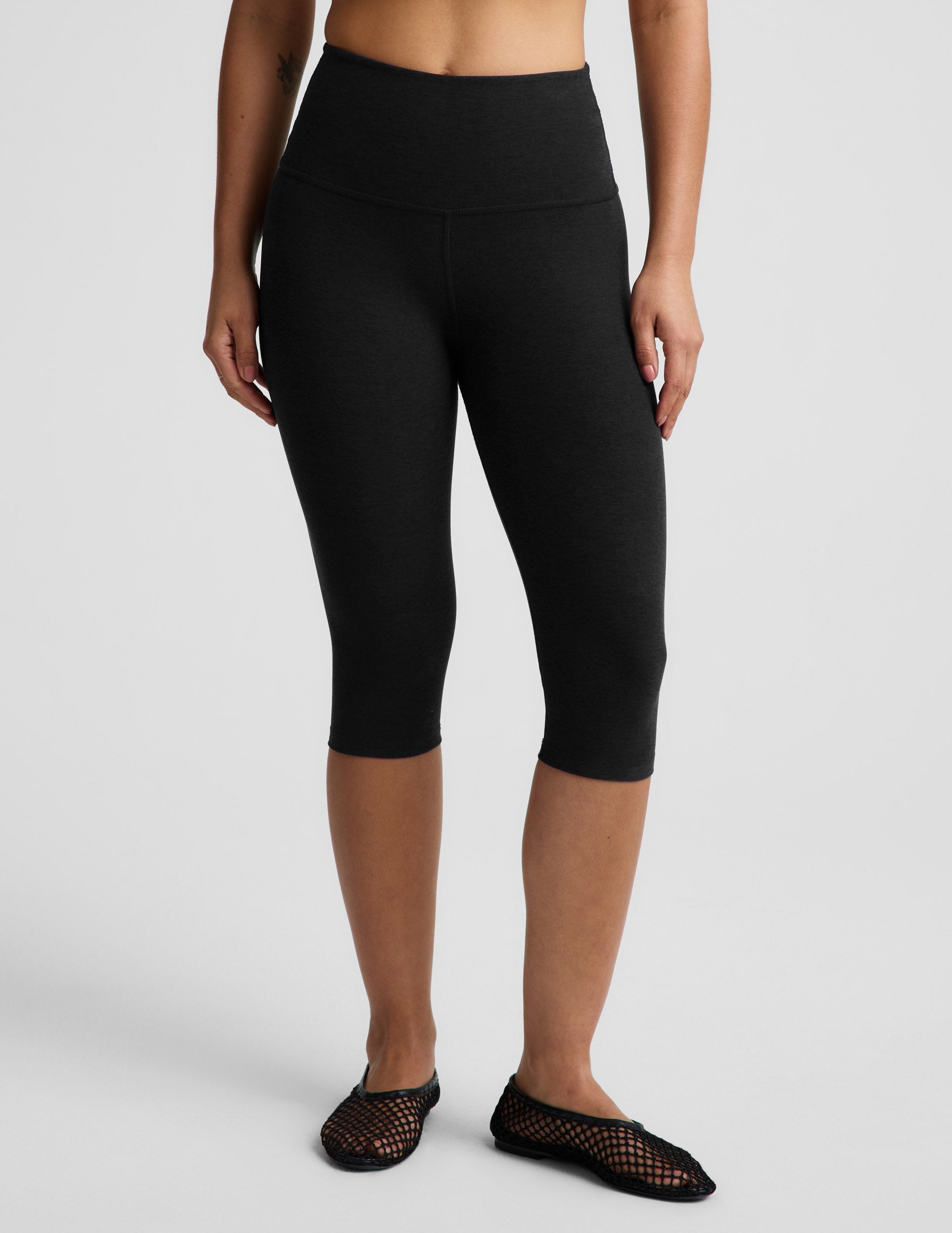 Woman wearing a white ribbed t-shirt and black pedal pusher leggings against a plain background.