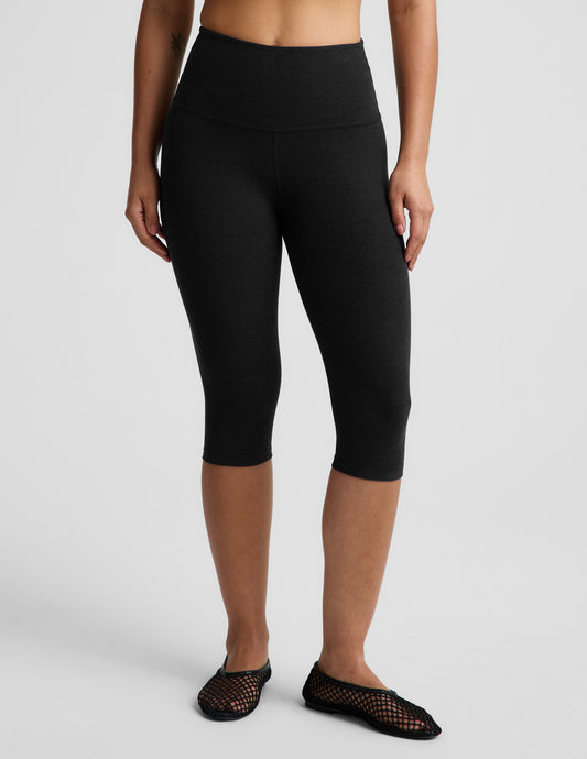 Woman wearing a white ribbed t-shirt and black pedal pusher leggings against a plain background.