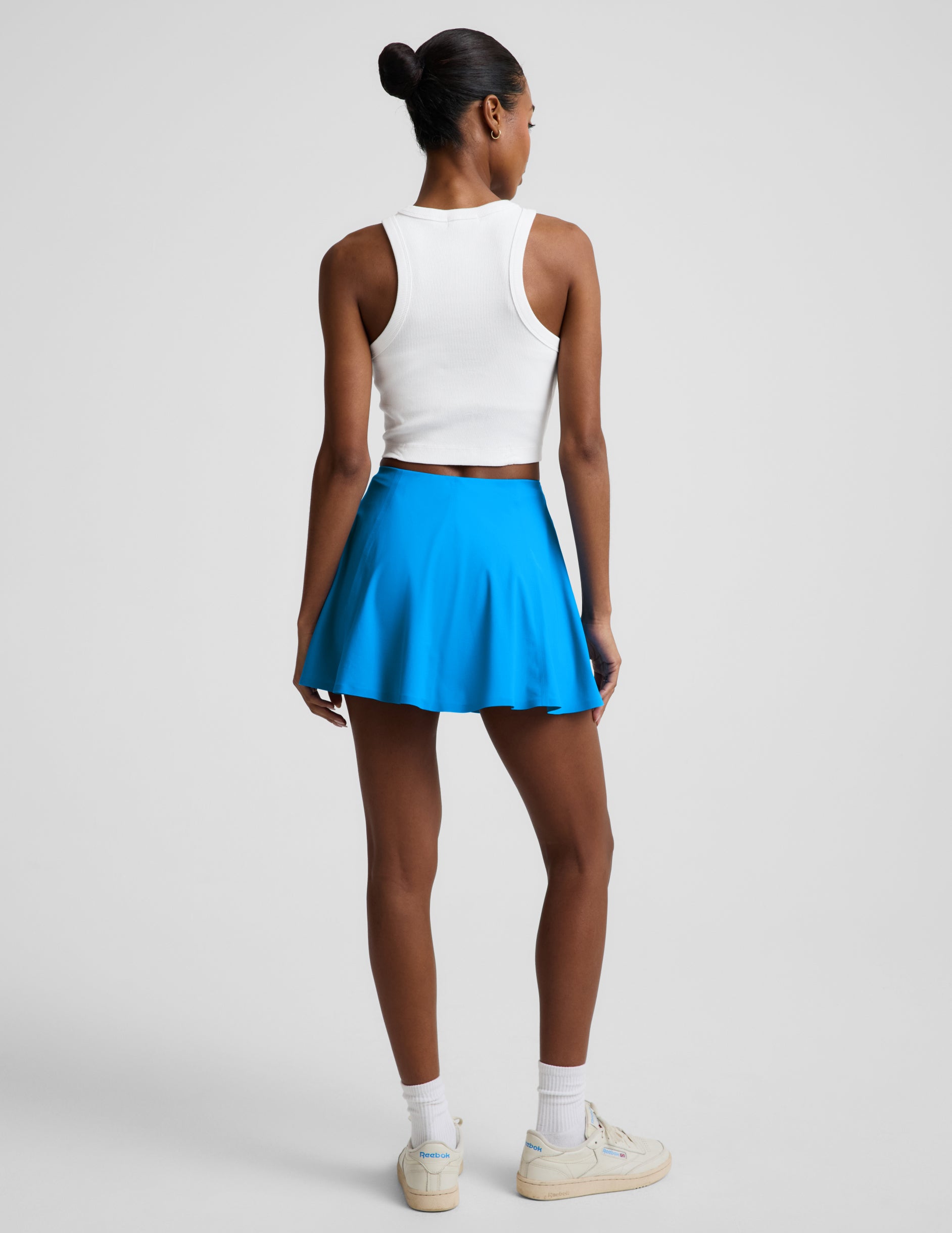 Rear view of woman wearing Blue You're A Force™ Skirt paired with a white tank top and cream sneakers.
