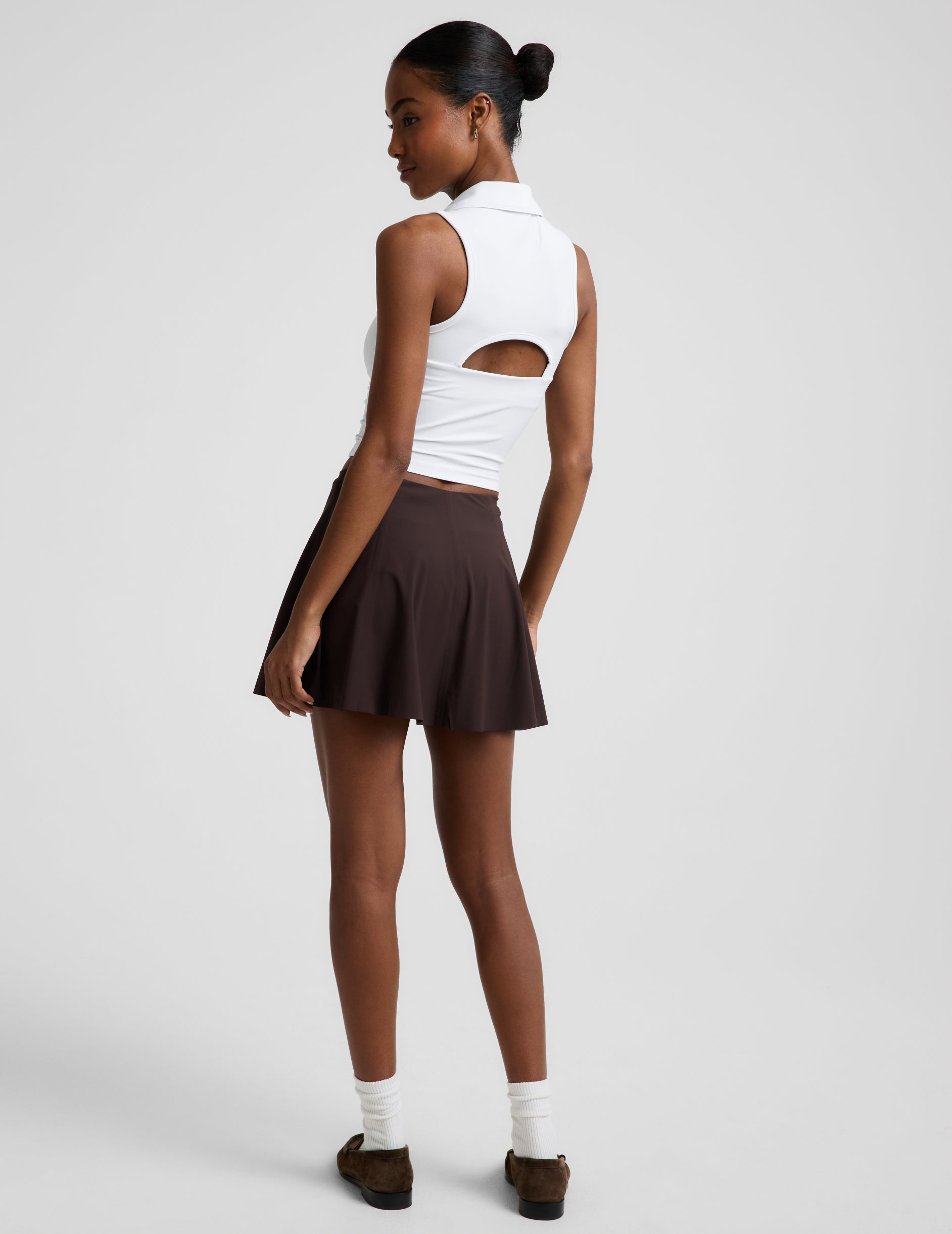 Woman wearing a white polo tank top and brown mini athletic skirt against a plain studio background.