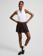 Woman wearing a white polo tank top and brown mini athletic skirt against a plain studio background.