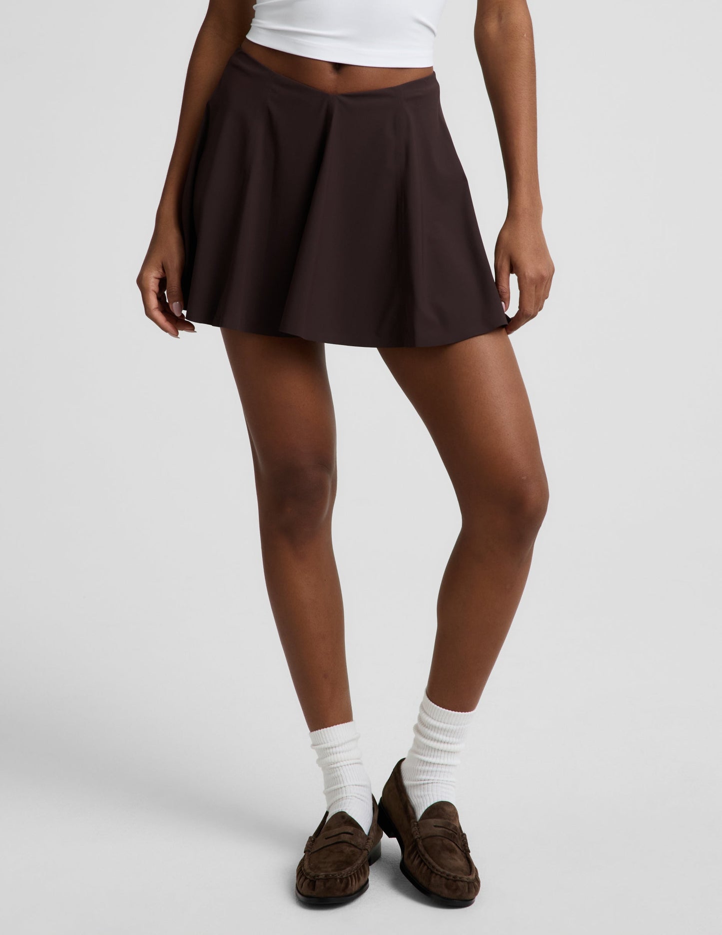 Woman wearing a white polo tank top and brown mini athletic skirt against a plain studio background.