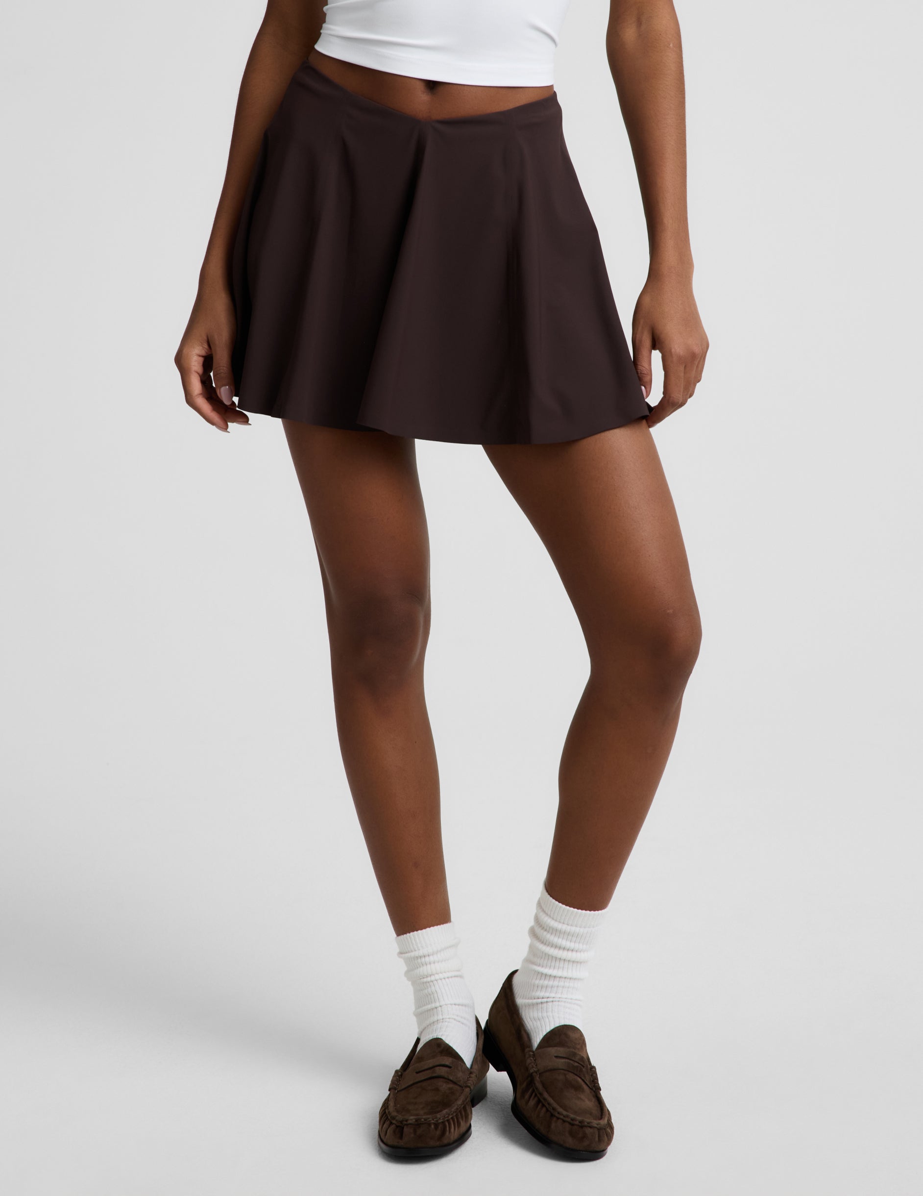 Woman wearing a white polo tank top and brown mini athletic skirt against a plain studio background.