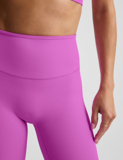 Woman wearing a purple sports bra and purple midi length leggings against a plain background.
