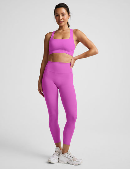 Woman wearing a purple sports bra and purple midi length leggings against a plain background.