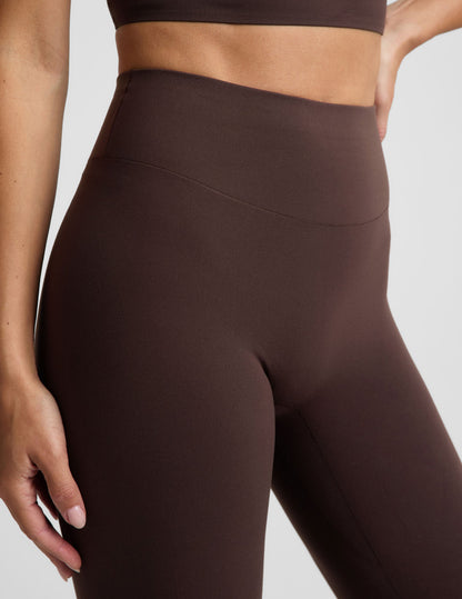 Woman wearing a brown cropped tank and brown midi length leggings against a plain studio background.