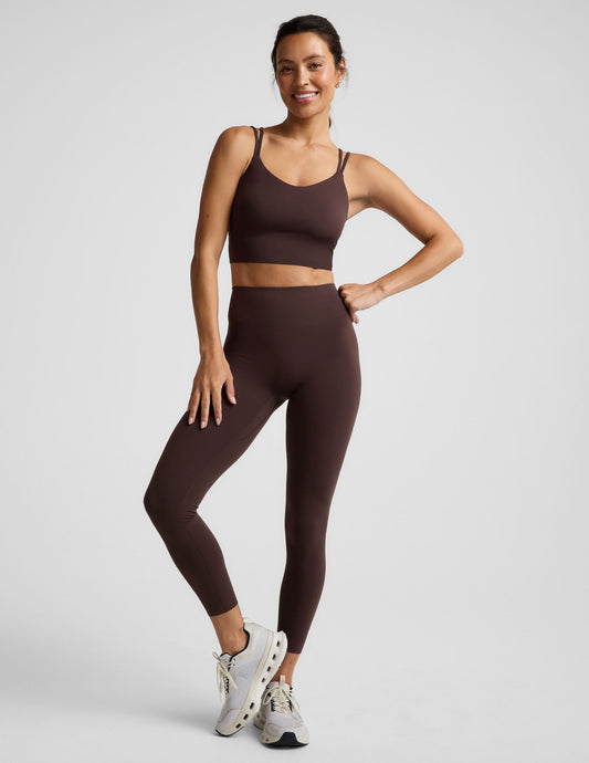 Woman wearing a brown cropped tank and brown midi length leggings against a plain studio background.