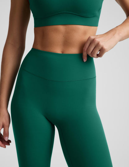 A close-up of a model wearing dark green athletic leggings and a matching sports bra against a light gray studio background.