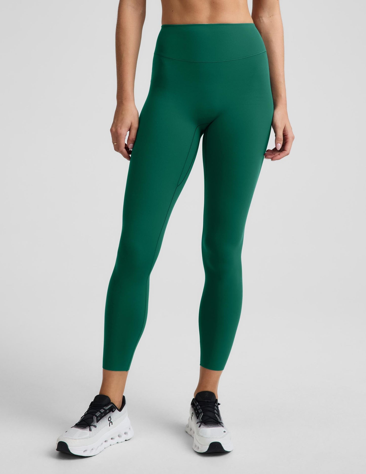 A model wearing high-waisted, full-length athletic leggings in a solid emerald green color, paired with white sneakers, shown from the waist down against a light gray studio background.