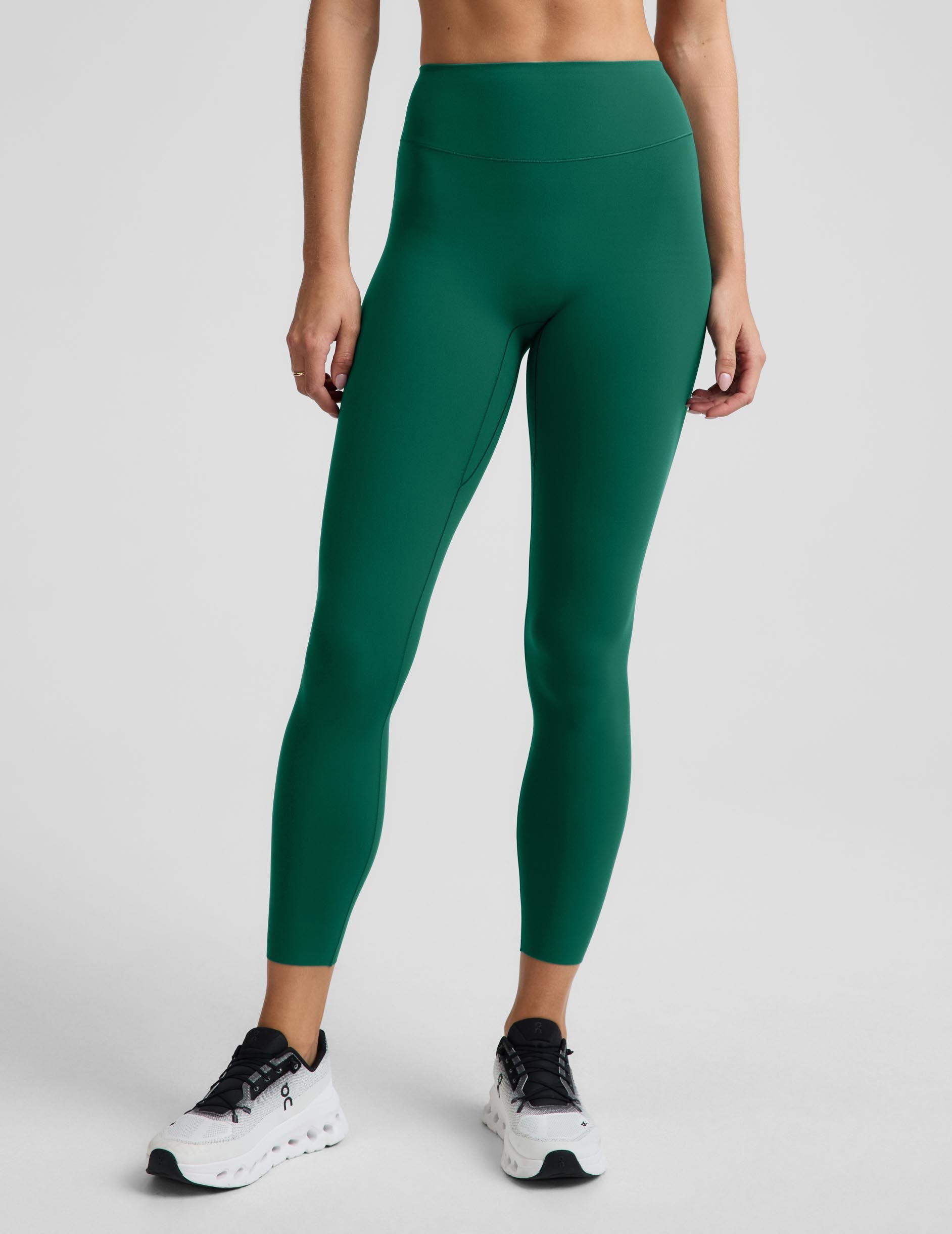 A model wearing high-waisted, full-length athletic leggings in a solid emerald green color, paired with white sneakers, shown from the waist down against a light gray studio background.