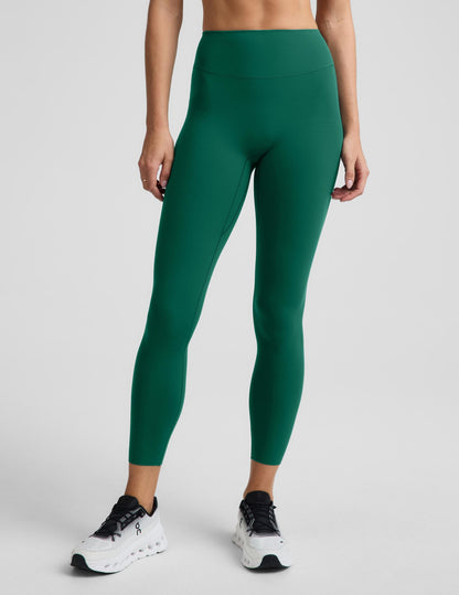 A model wearing high-waisted, full-length athletic leggings in a solid emerald green color, paired with white sneakers, shown from the waist down against a light gray studio background.