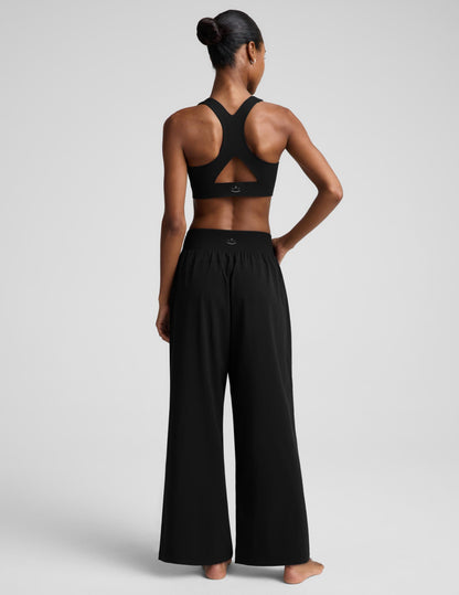 A woman modeling a black two-piece activewear set consisting of a cropped tank top and high-waisted wide-leg pants, photographed against a solid light gray studio background.
