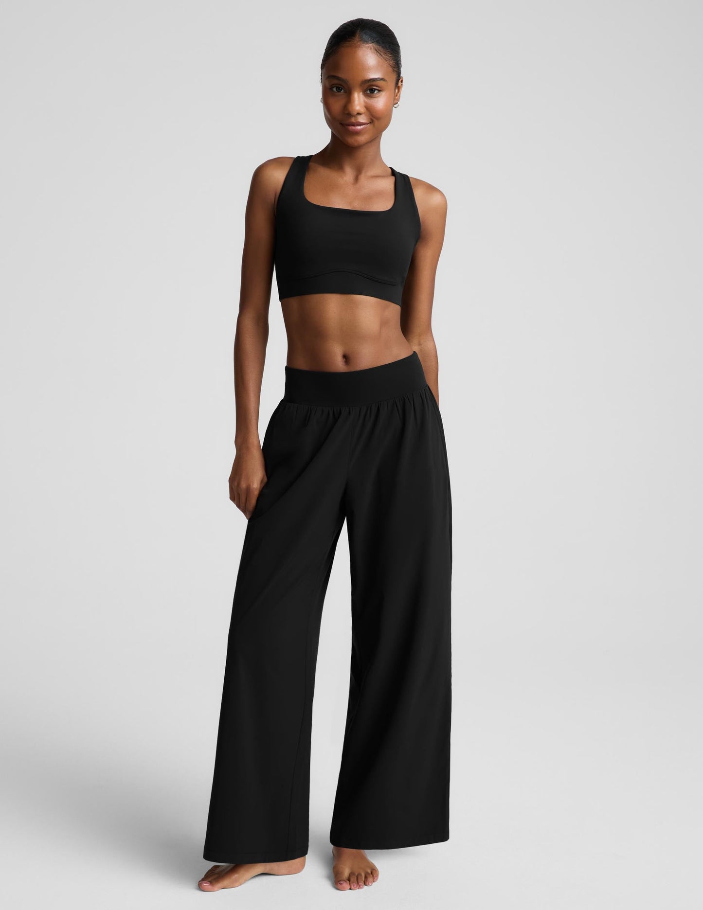 A woman modeling a black two-piece activewear set consisting of a bra and high-waisted wide-leg pants, photographed against a solid light gray studio background.
