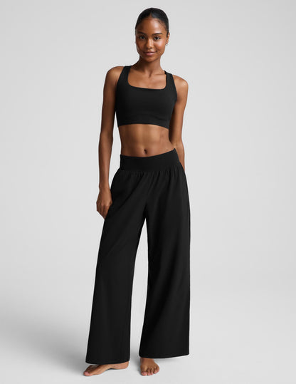 A woman modeling a black two-piece activewear set consisting of a bra and high-waisted wide-leg pants, photographed against a solid light gray studio background.