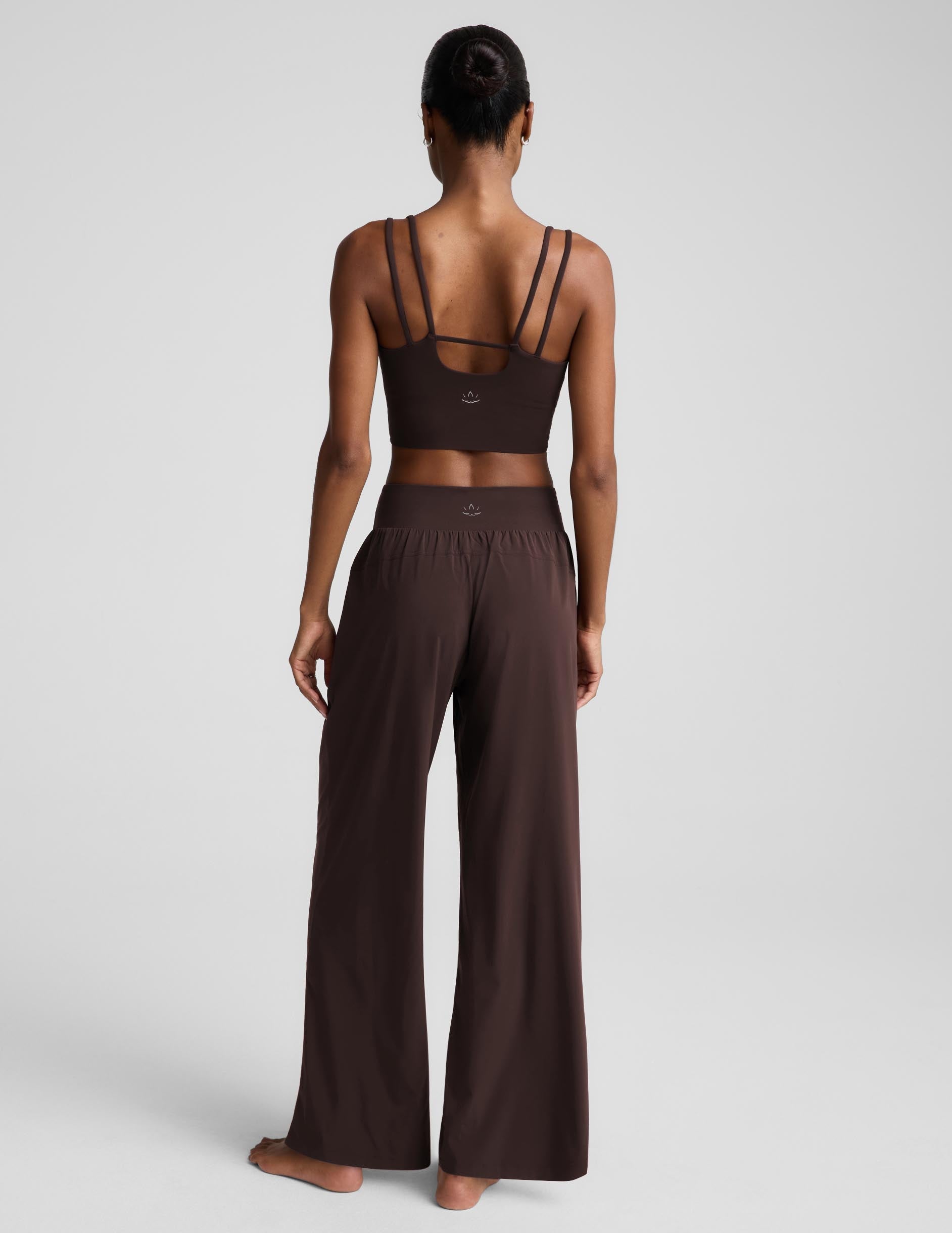 A woman modeling a matching dark brown lounge set consisting of a cropped tank top and wide-leg pants, photographed against a neutral light gray studio background.