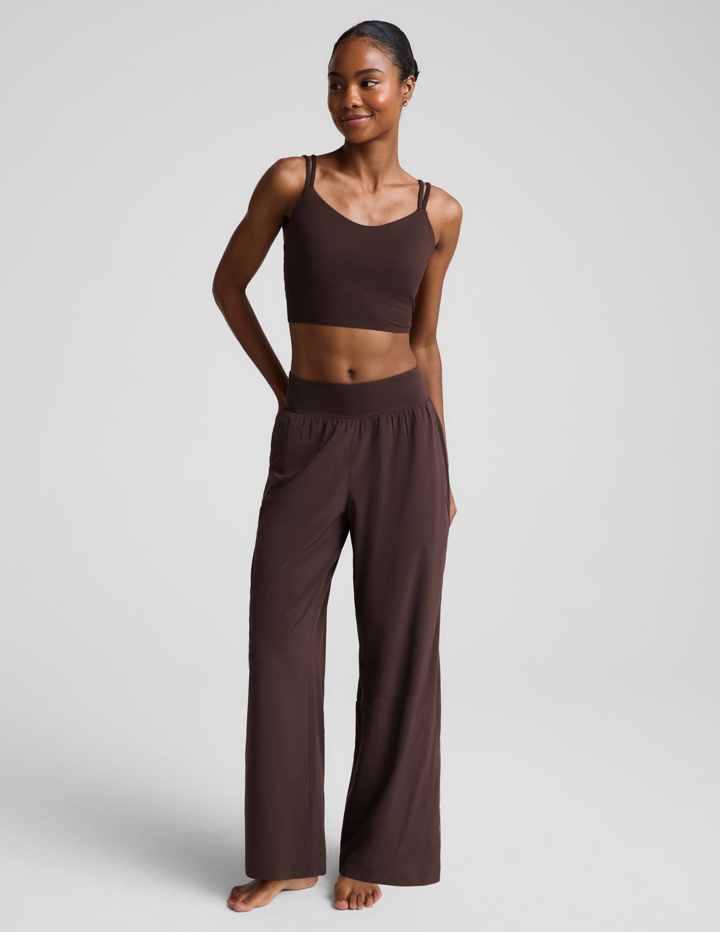 A woman modeling a matching dark brown lounge set consisting of a cropped tank top and wide-leg pants, photographed against a neutral light gray studio background.