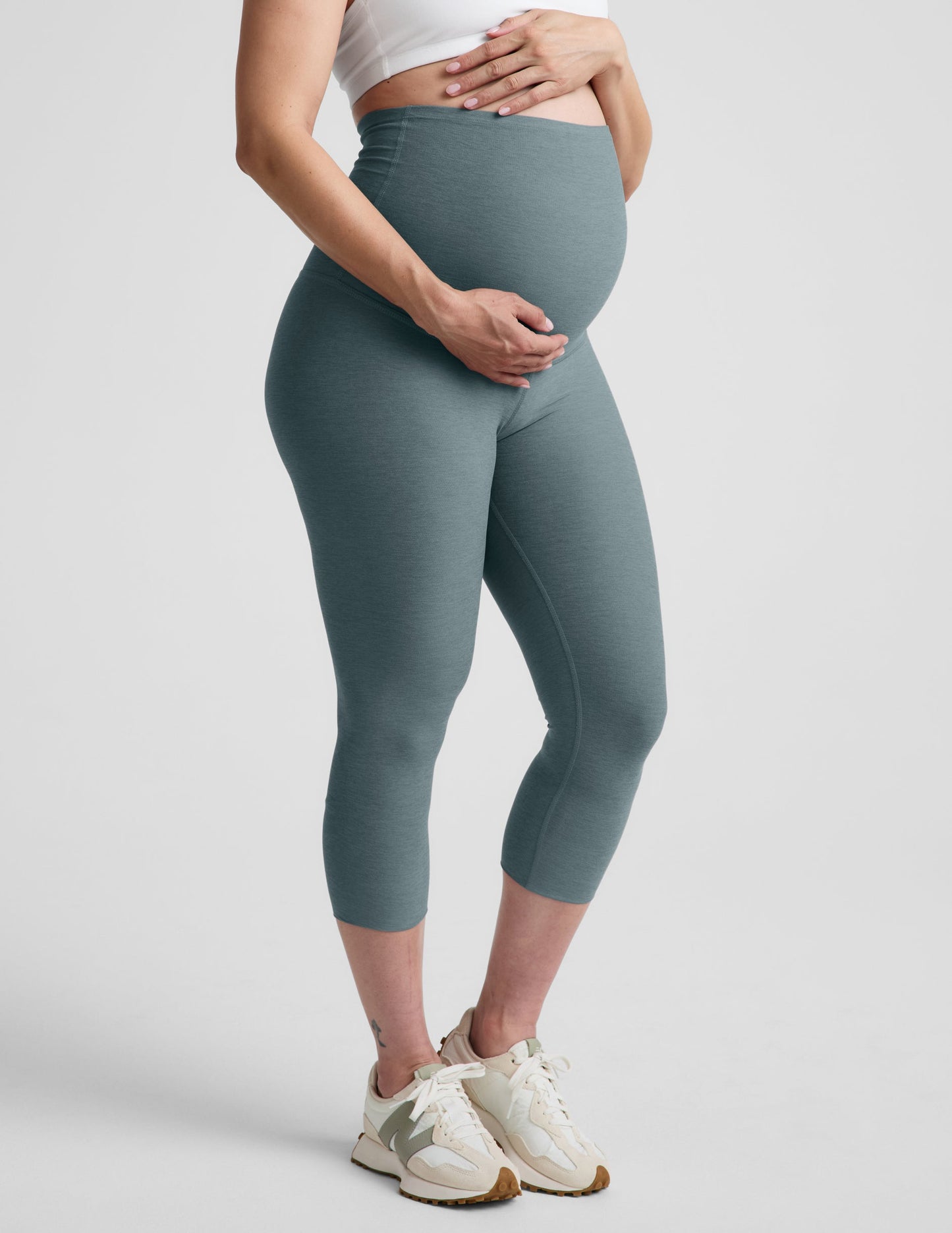 Woman wearing Blue Spacedye™ Love The Bump Maternity 20.5" Capri Legging with white sports bra and cream sneakers. Side view.