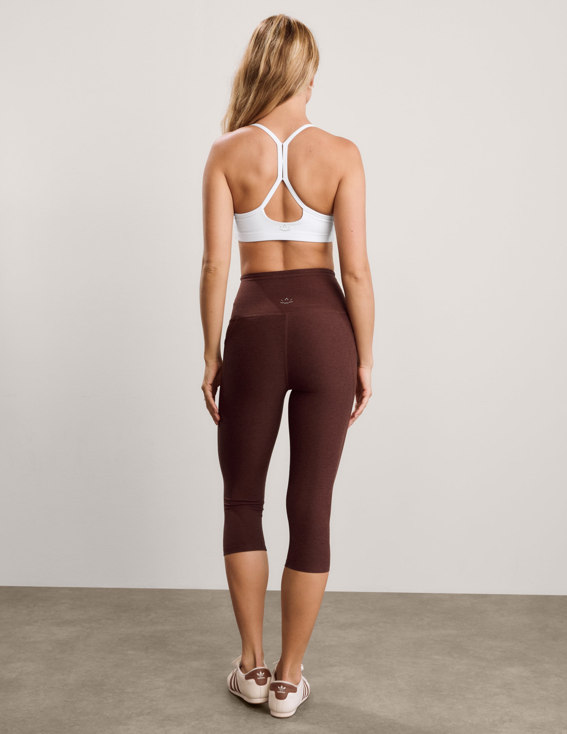 Woman wearing a white sports bra and brown capris with pockets against a plain background.