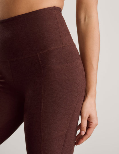 Woman wearing a white sports bra and brown capris with pockets against a plain background.