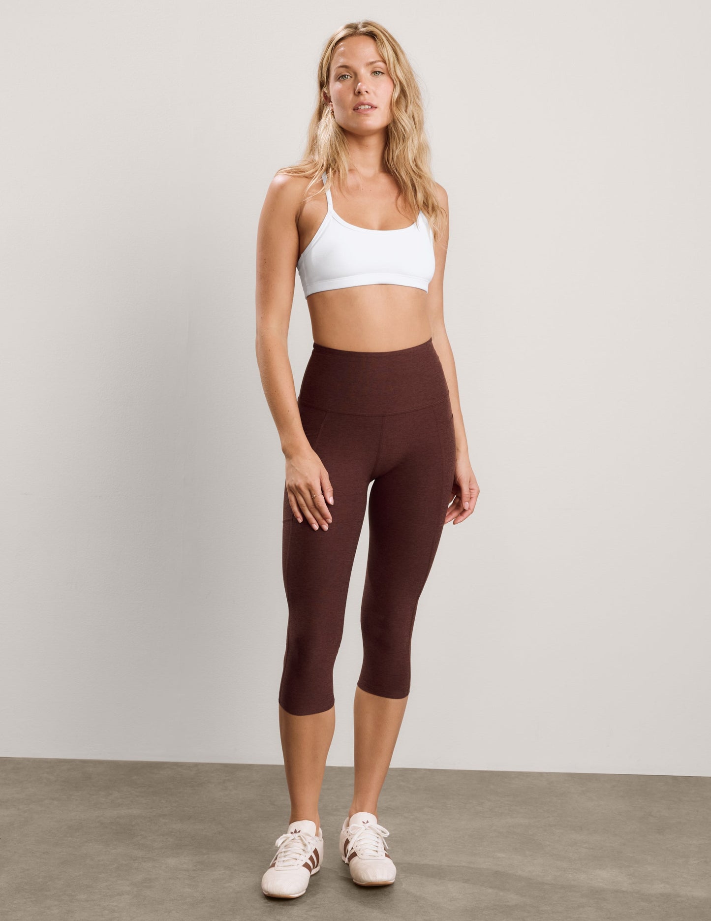 Woman wearing a white sports bra and brown capris with pockets against a plain background.