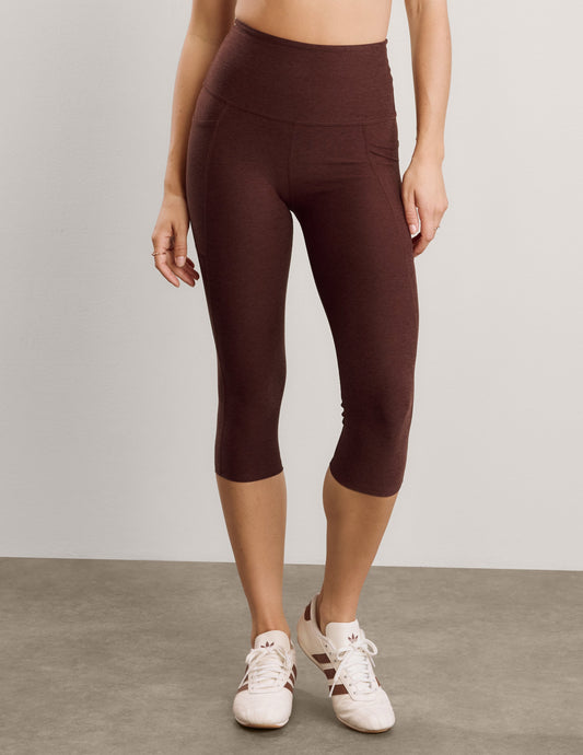 Woman wearing a white sports bra and brown capris with pockets against a plain background.