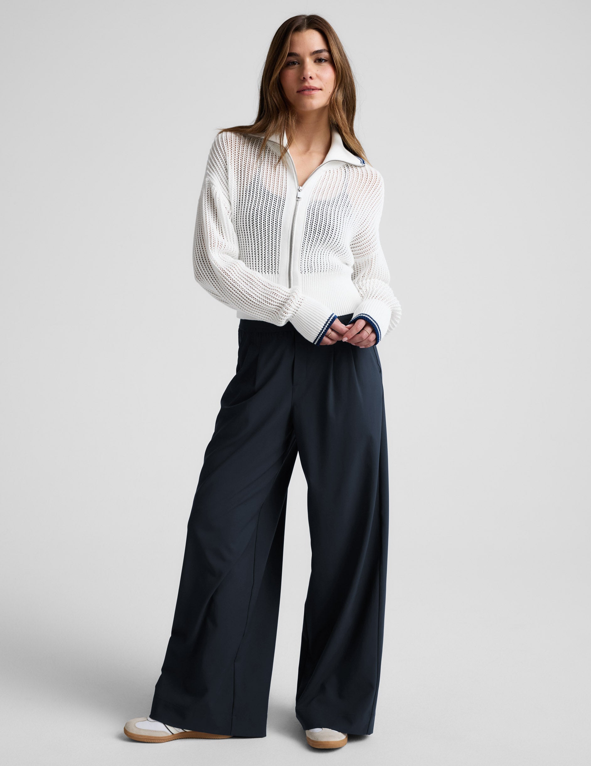 Woman wearing a white zip-up sweater and blue trousers against a plain background.