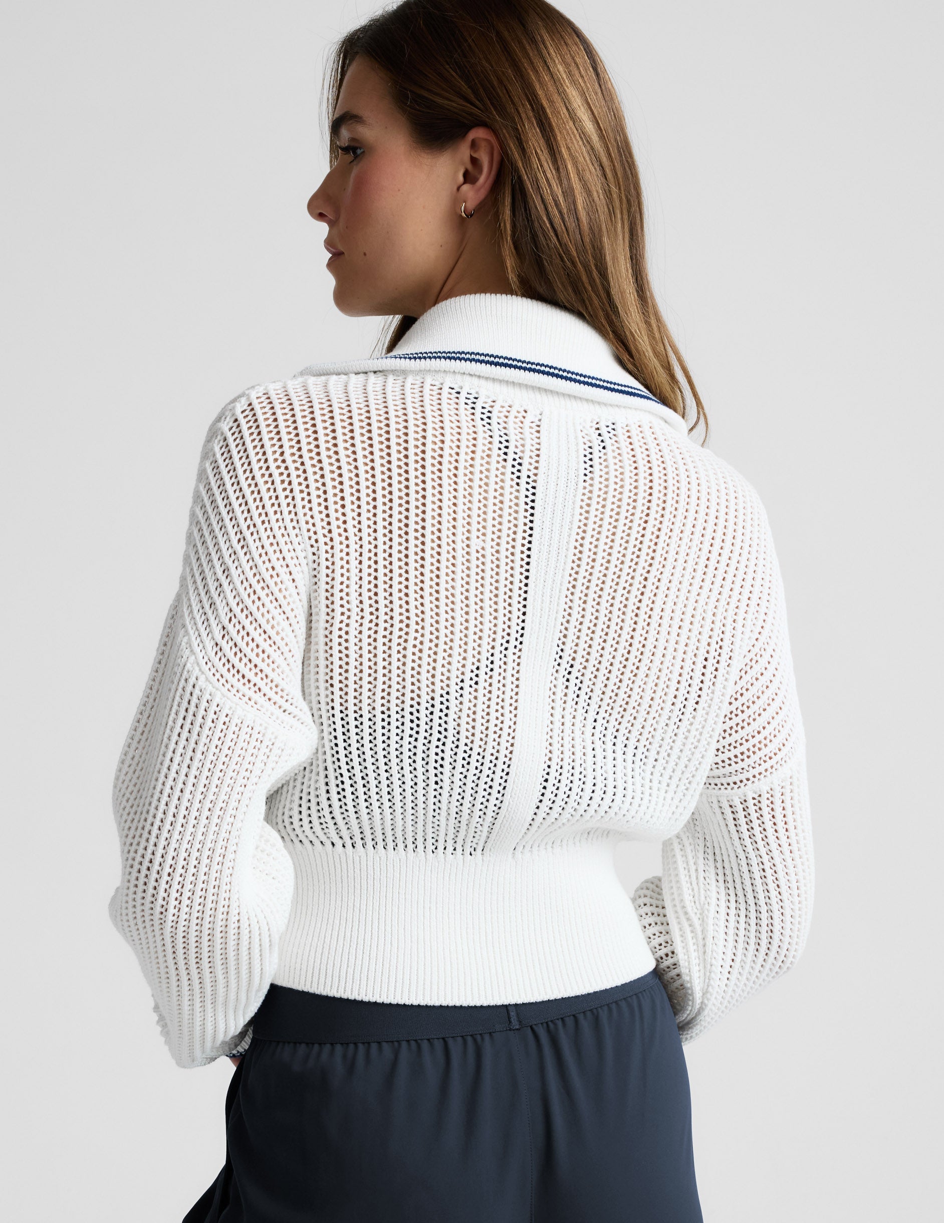 Woman wearing a white zip-up sweater and blue trousers against a plain background.