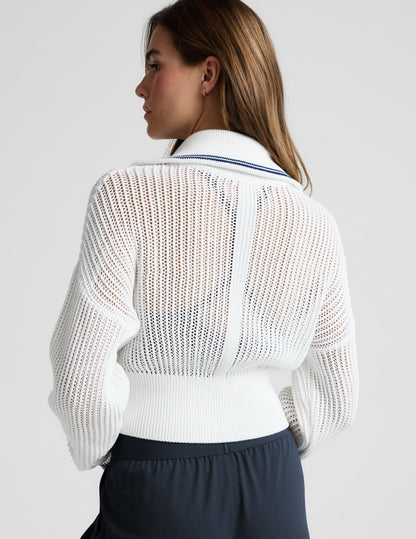 Woman wearing a white zip-up sweater and blue trousers against a plain background.