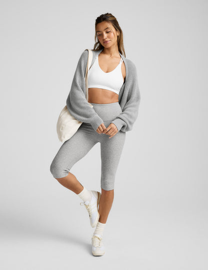 Woman wearing a gray shrug, white sports bra, and gray leggings on a light gray background.