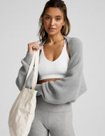 Woman wearing a gray shrug, white sports bra, and gray leggings on a light gray background.