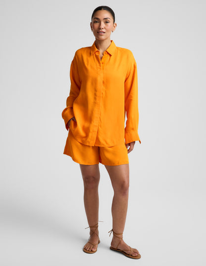 Woman wearing an orange button down shirt and orange shorts against a plain background.