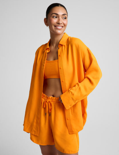 Woman wearing an orange button down shirt and orange shorts against a plain background.