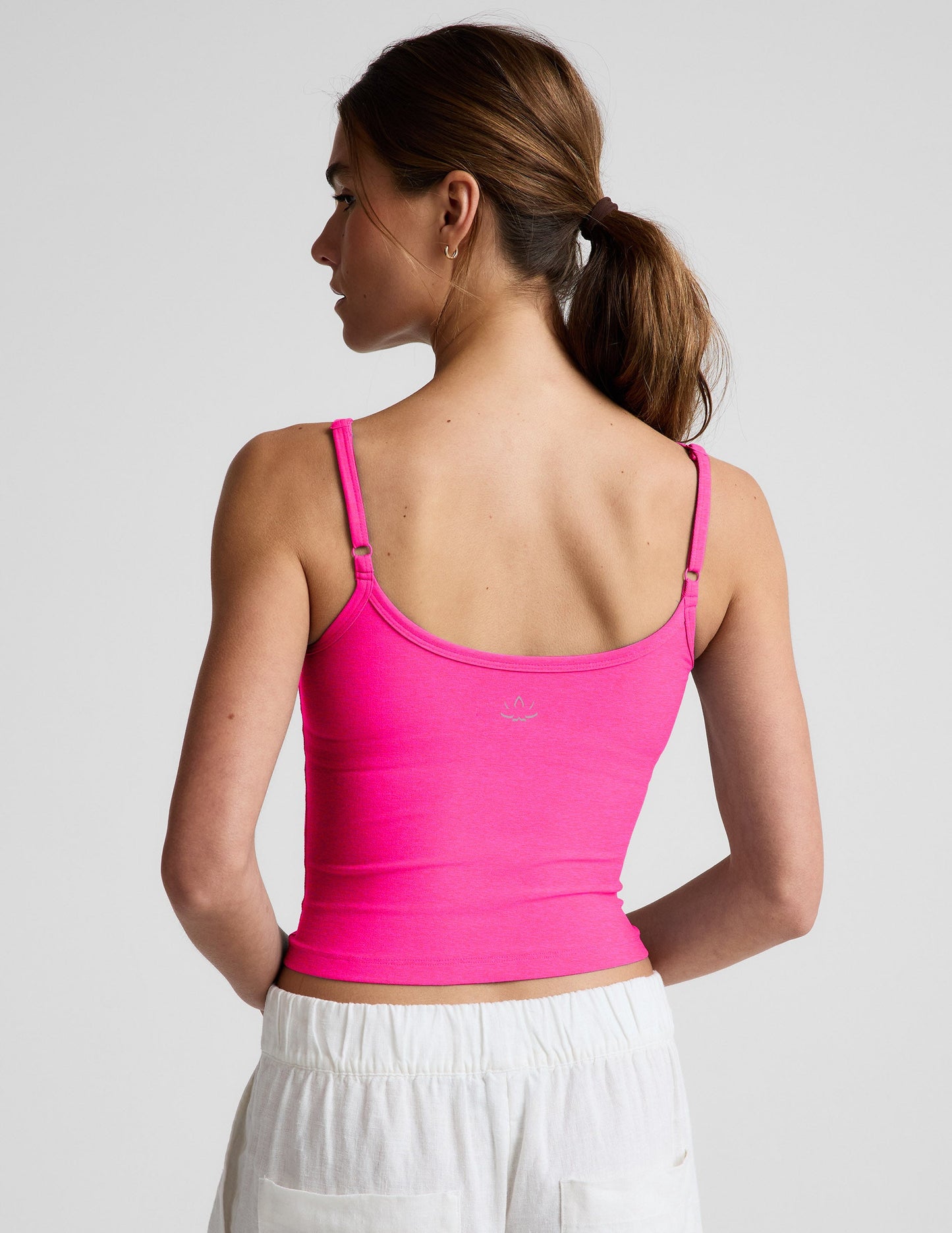 Woman wearing a bright pink waist length tank top and white linen pants against a plain background.