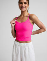 Woman wearing a bright pink waist length tank top and white linen pants against a plain background.