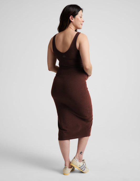 Pregnant woman wearing a brown maternity midi length dress with a front slit against a plain background.