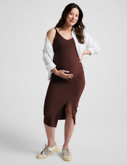 Pregnant woman wearing a brown maternity midi length dress with a front slit against and a white poplin shirt a plain background.
