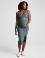 Pregnant woman wearing a green maternity midi length tank dress with a front side slit against a plain background.