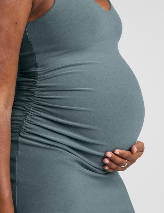 Pregnant woman wearing a green maternity midi length tank dress with a front side slit against a plain background.