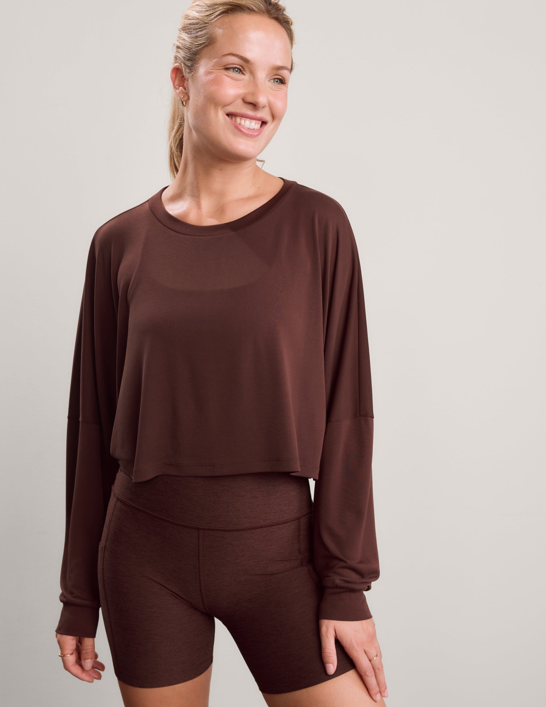 Woman wearing a brown long sleeve top and brown biker shorts against a plain background.